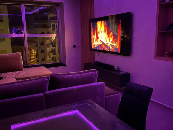 Salon d'un appartement de luxe à Tanger Marina Bay avec canapé en velours, éclairage néon violet, TV écran plat et vue sur la ville de nuit.