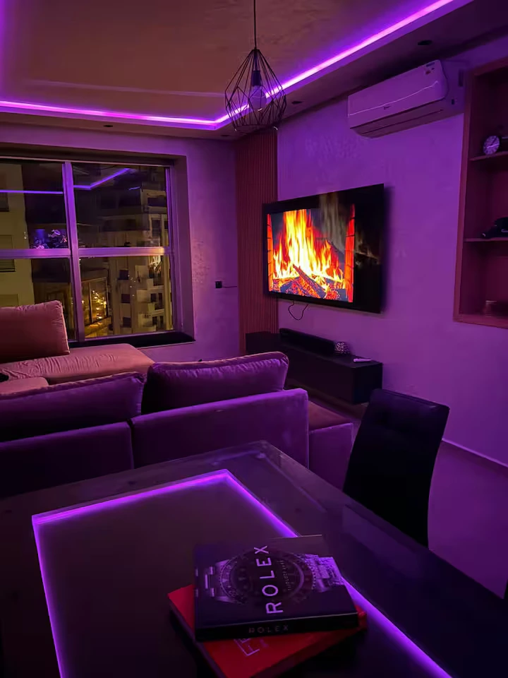 Salon d'un appartement de luxe à Tanger Marina Bay avec canapé en velours, éclairage néon violet, TV écran plat et vue sur la ville de nuit.