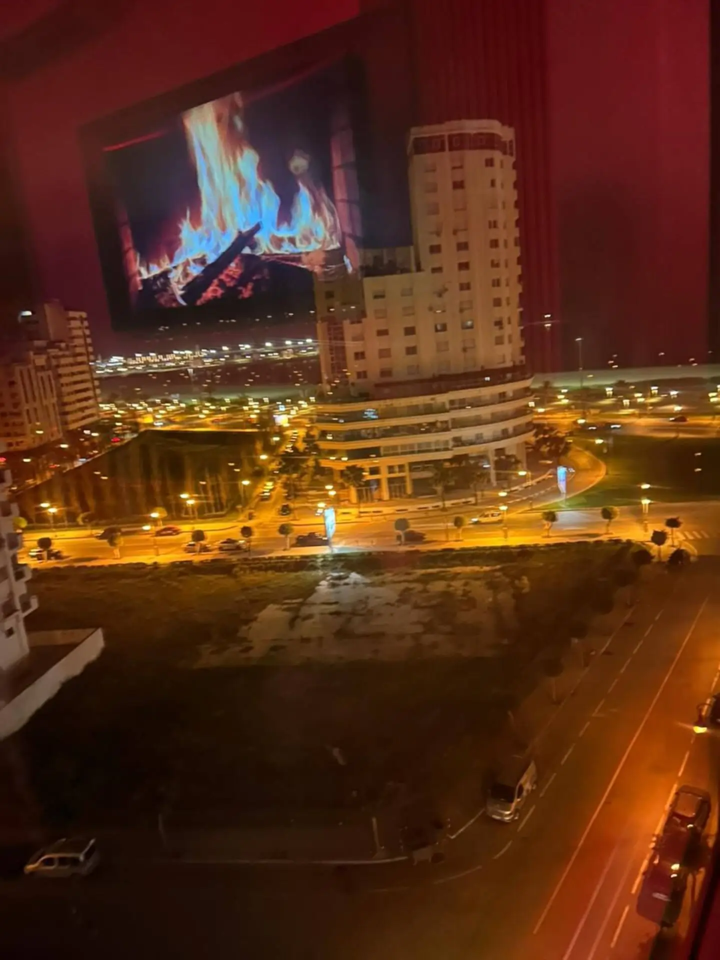 vue-appartement-luxe-nuit-ville-moderne Vue panoramique nocturne depuis un appartement de haut standing surplombant un boulevard illuminé et une architecture urbaine moderne, avec un reflet chaleureux de cheminée en intérieur.